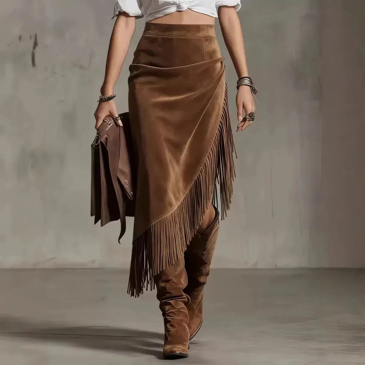 Modelo caminhando com saia midi de camurça marrom com franjas, camisa branca e bota, em parede de concreto