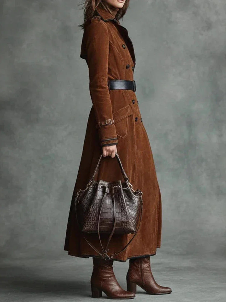 Modelo de trench coat de camurça marrom com cinto preto e bolsa de couro, em fundo cinza esverdeado