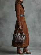 Modelo de trench coat de camurça marrom com cinto preto e bolsa de couro, em fundo cinza esverdeado